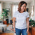 Frau in weißem T Shirt und Jeans steht lächelnd in einem hellen Wohnzimmer mit Sofa Teppich und mehreren Zimmerpflanzen im Hintergrund. Die Szene vermittelt eine entspannte Alltagssituation in einem gemütlich eingerichteten Zuhause.